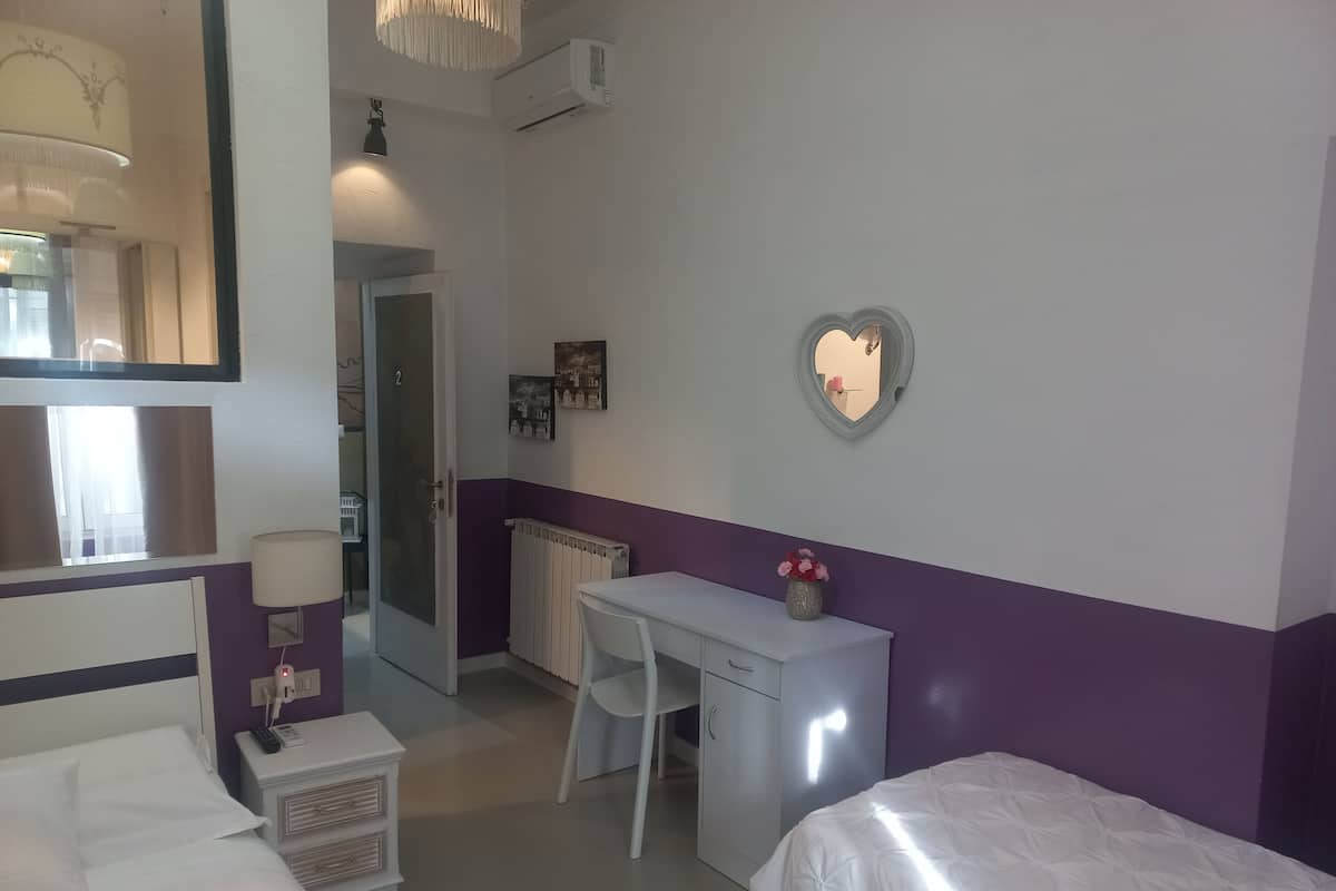A hotel room with a bed, a desk, a chair, a small bedside table, a mirror, and a radiator.