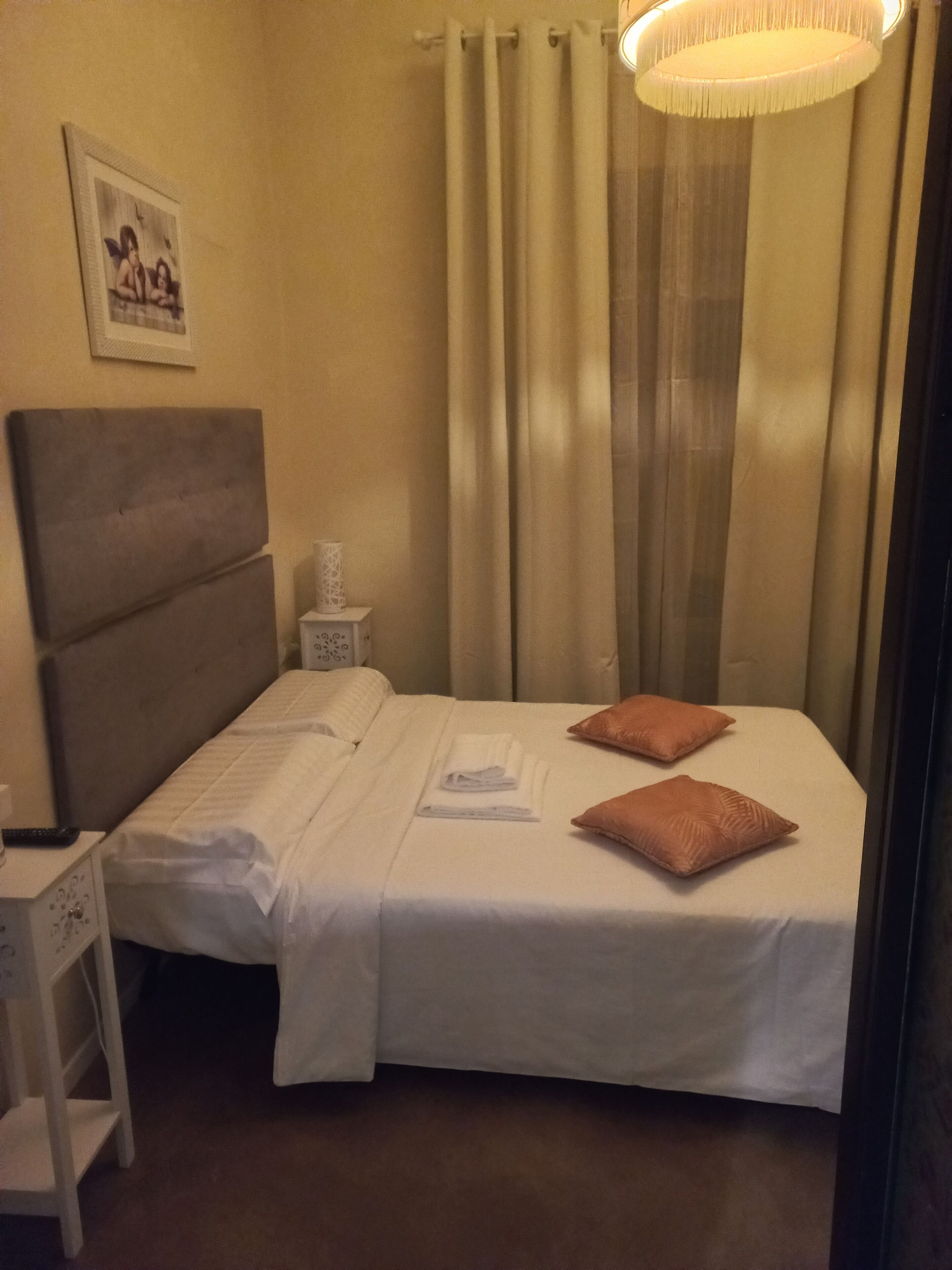 A neatly made bed with white linens and two brown pillows, a bedside table with a lamp, and a framed picture on the wall.