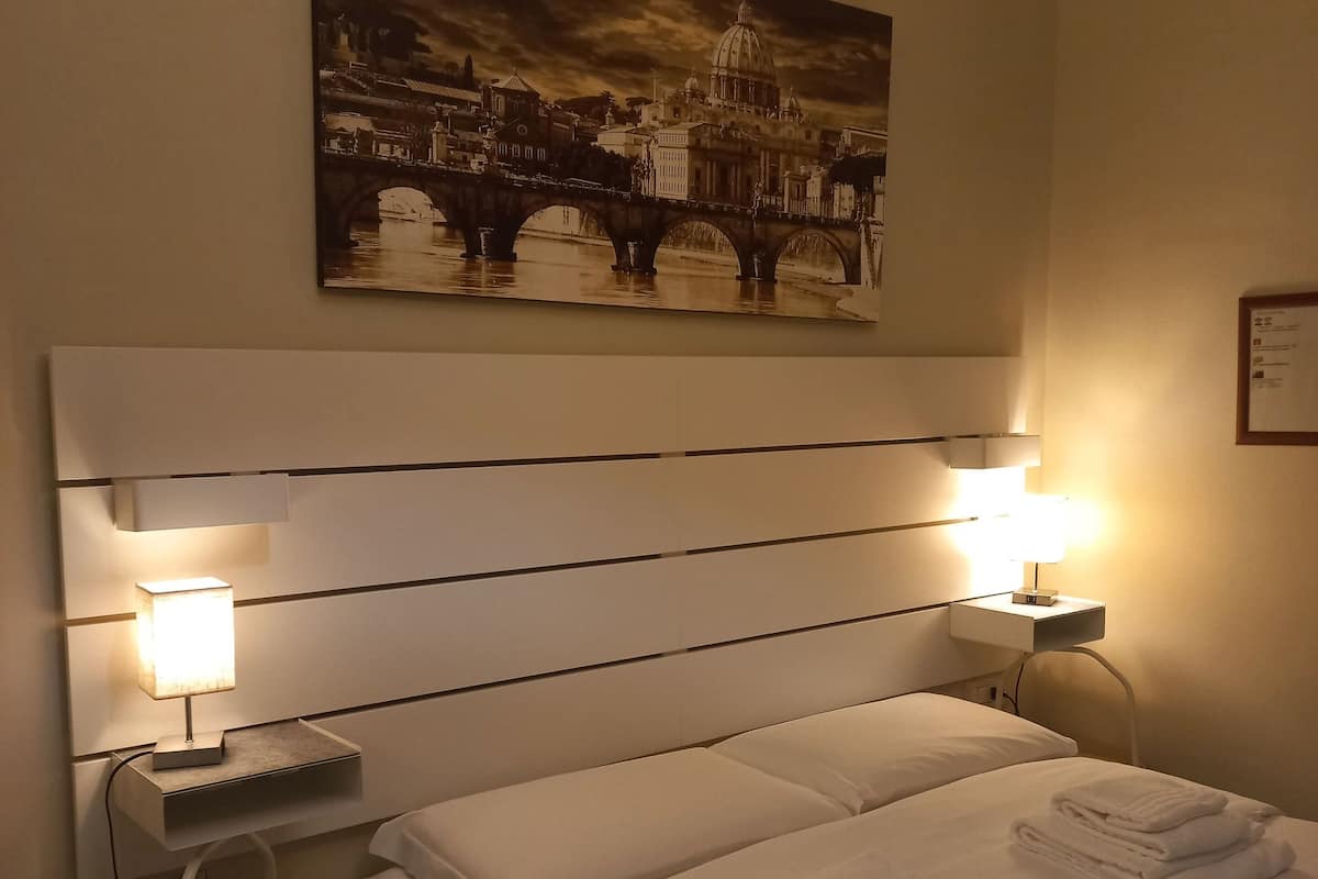 A hotel room with a large bed, two bedside lamps, a small table with a remote control, and a painting of a cityscape on the wall.