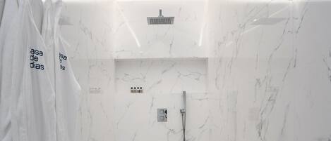 Un baño moderno con paredes de mármol blanco, ducha con cabezal de ducha de mano y un pequeño estante con artículos de aseo.