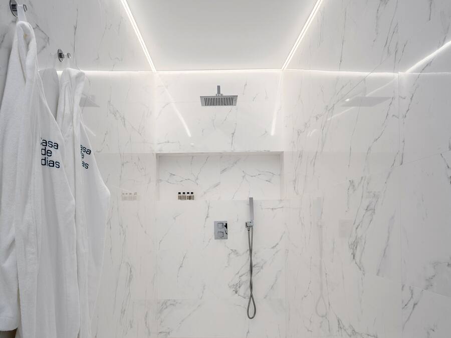 A modern bathroom with white marble walls, a shower with a handheld showerhead, and a small shelf with toiletries.
