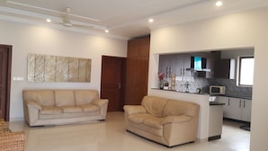 A living room with a beige sofa, a ceiling fan, and a kitchen area with a microwave and cabinets. - Valley View Villa (Rawalpindi)