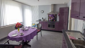 Ett modernt kök med lila skåp, ett matbord med en lila duk och ett stort fönster med persienner.