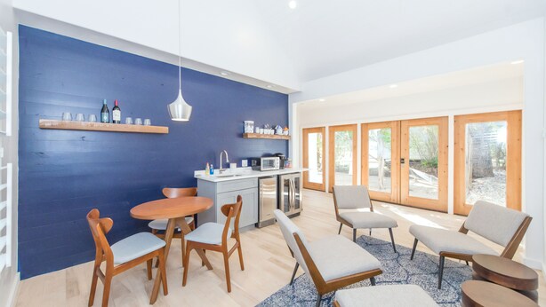 A modern living room with a blue accent wall, wooden furniture, a kitchen area with a microwave, and large windows. - Benson Guest House (Uvalde)