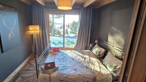 A bedroom with a bed, a bedside table, a lamp, and a window with a view of a snowy landscape. - Chalet lovers in Switzerland (Saint George)