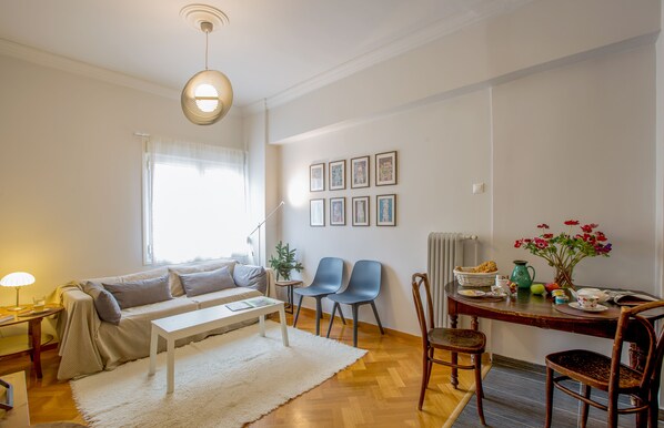 Een gezellige woonkamer met een bank, een salontafel en een eettafel met stoelen. Er hangen afbeeldingen in lijsten aan de muur, een plant en een vaas met bloemen op de eettafel.