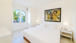 A bedroom with a large bed, two bedside tables with lamps, a window with a view of palm trees, and a painting on the wall. - CAN RAMIS -  Seafront house with panoramic views. (Cielo de Bonaire)