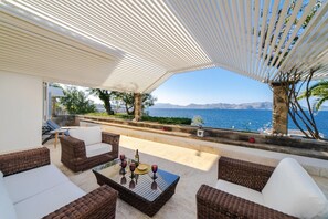A sunlit outdoor seating area with wicker furniture and a glass table, overlooking a scenic coastal view.