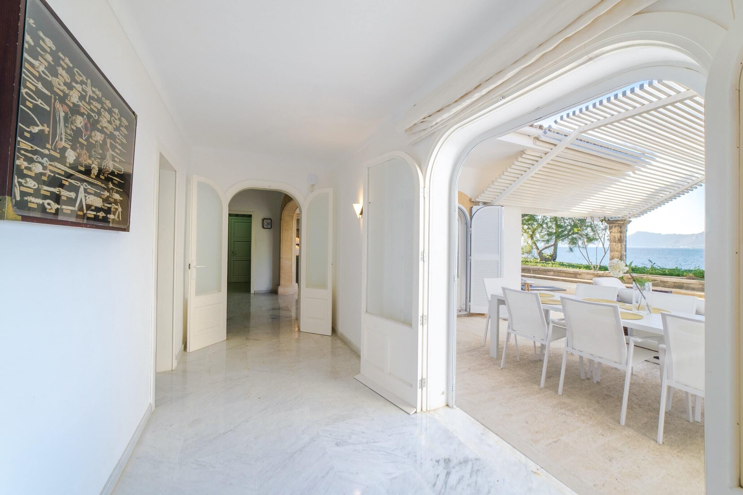 A corridor with white walls and a marble floor leading to a covered outdoor seating area with white furniture.
