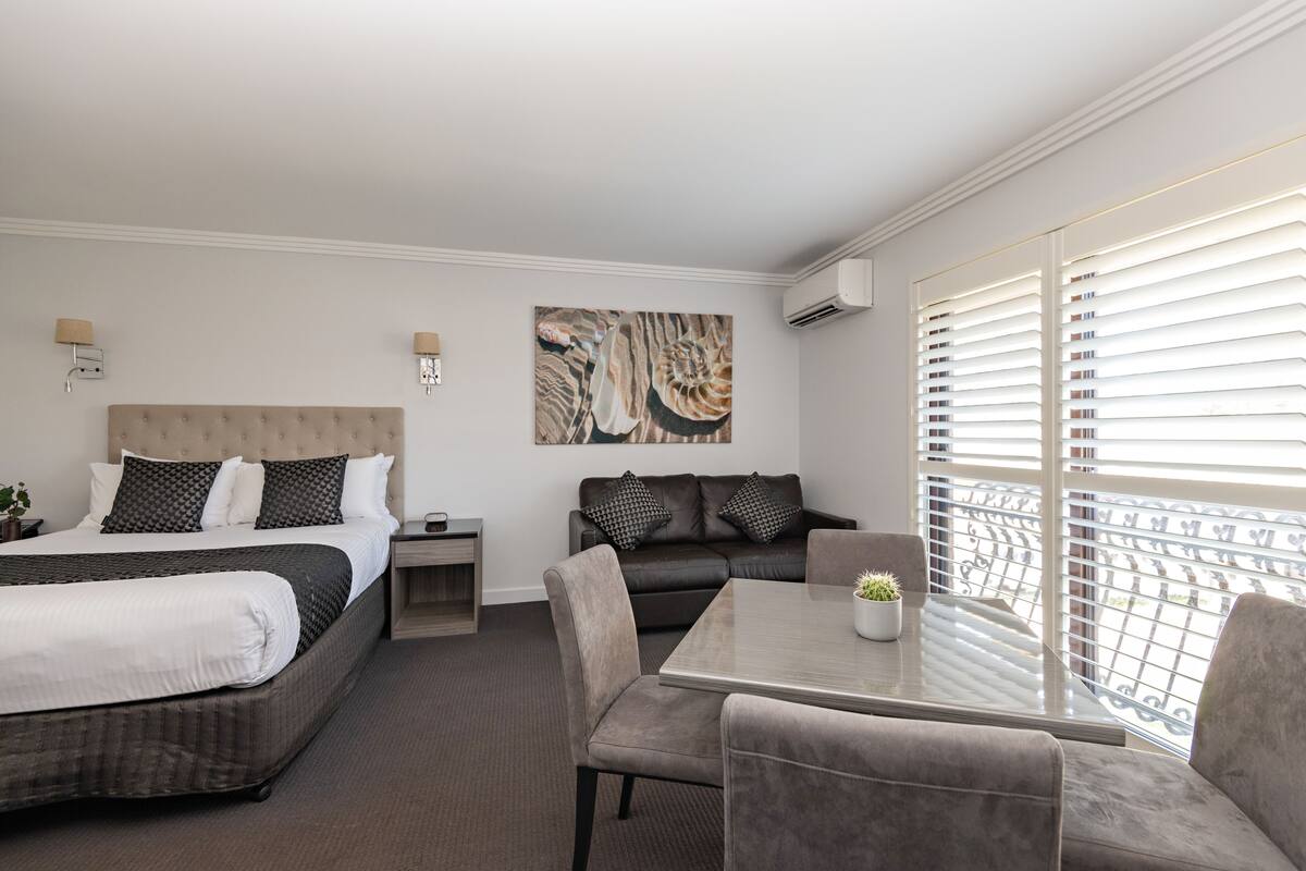 Una habitación de hotel con cama, sofá, mesa de comedor y un ventanal con persianas.