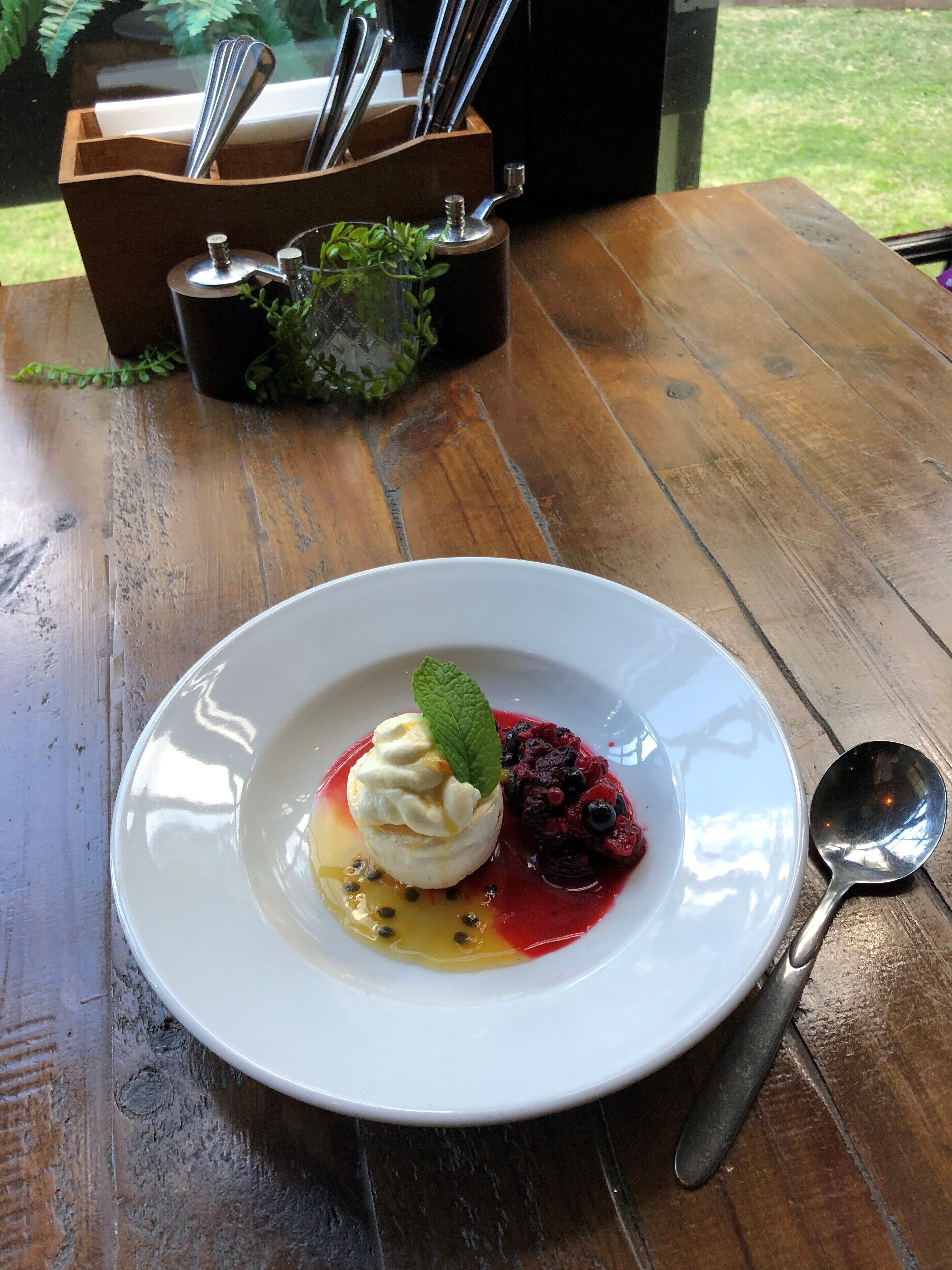 A dessert on a white plate with a mint leaf on top, accompanied by berries and a sauce, on a wooden table with a spoon beside it.