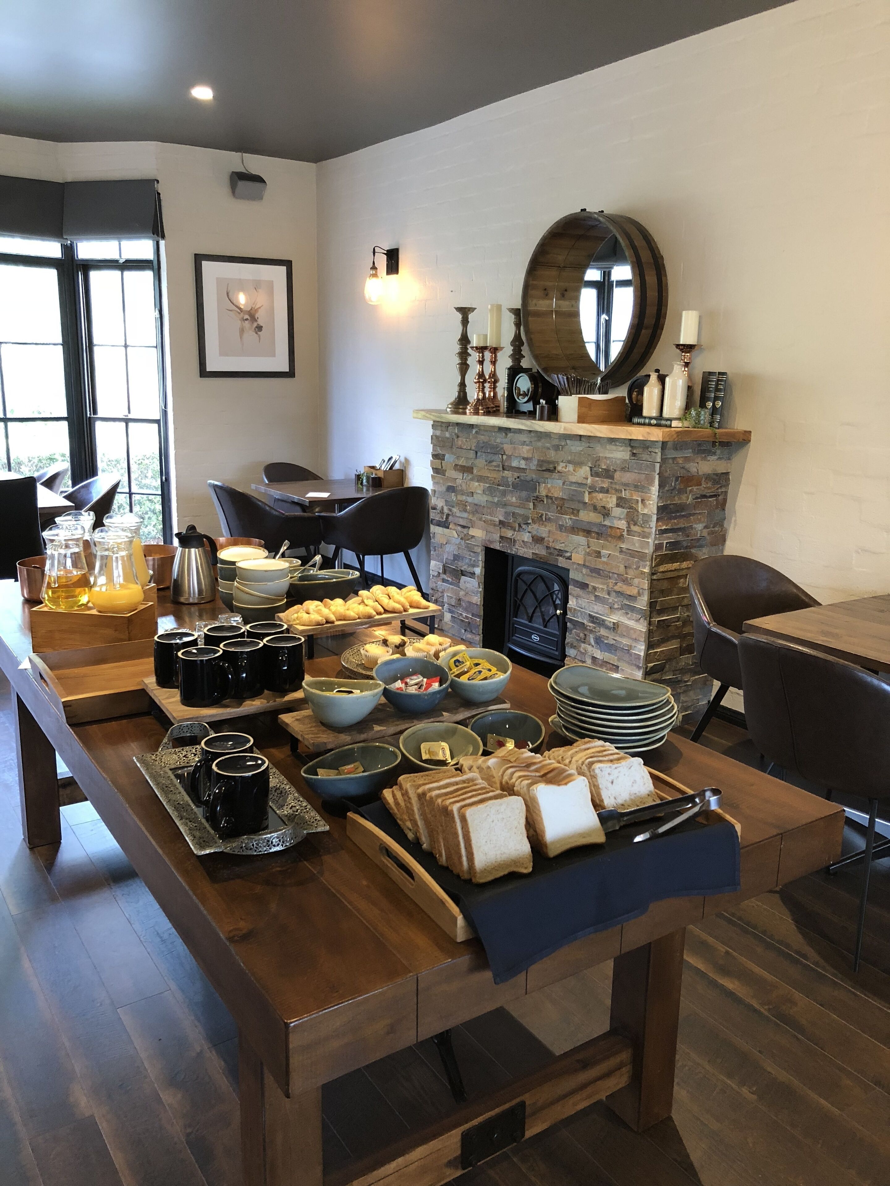 A breakfast buffet with various food items and beverages, a stone fireplace, and modern furniture in a well-lit room.
