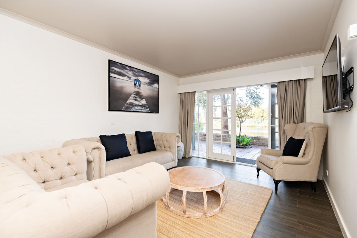 Una sala de estar con un sofá color crema, una mesa de centro redonda de madera y un sillón color crema. Hay un ventanal con cortinas, un televisor de pantalla plana montado en la pared y un cuadro sobre el sofá.