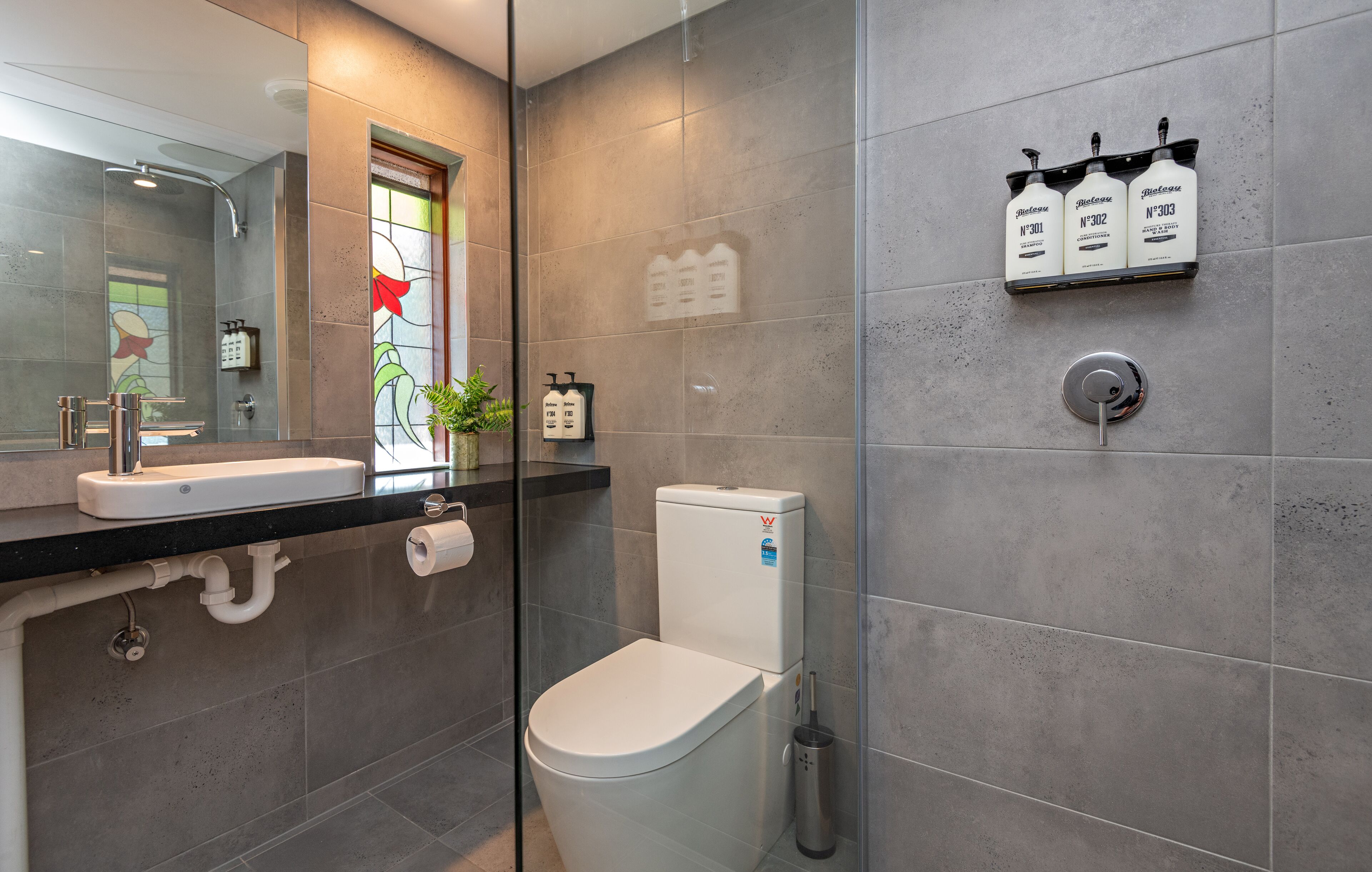 A modern bathroom with a glass shower enclosure, a toilet, a sink with a mirror, and a shelf with toiletries.
