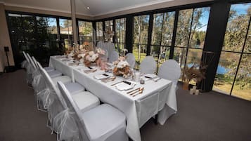 A long dining table set with white chairs, floral centerpieces, and a view of a lake through large windows.