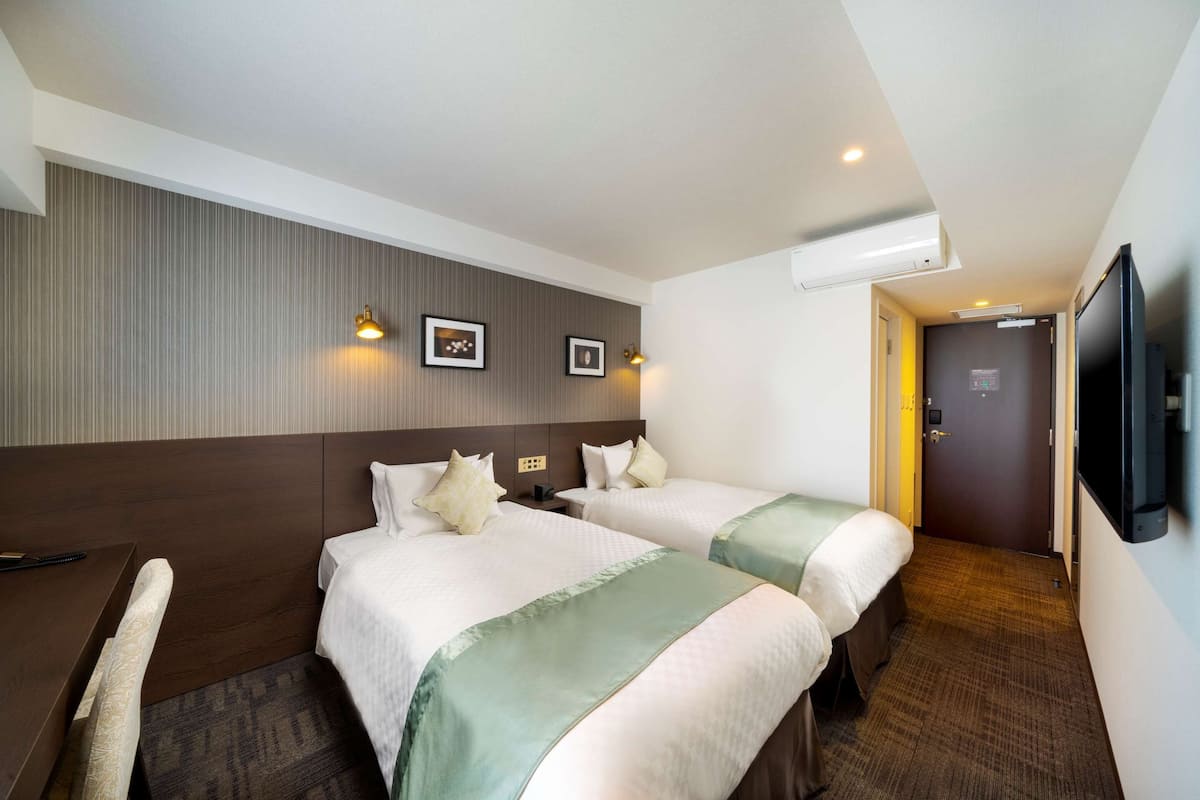 Ein Hotelzimmer mit zwei Betten, einem Schreibtisch, einem Sessel, einem Flachbildfernseher und einer Tür zu einem weiteren Raum.