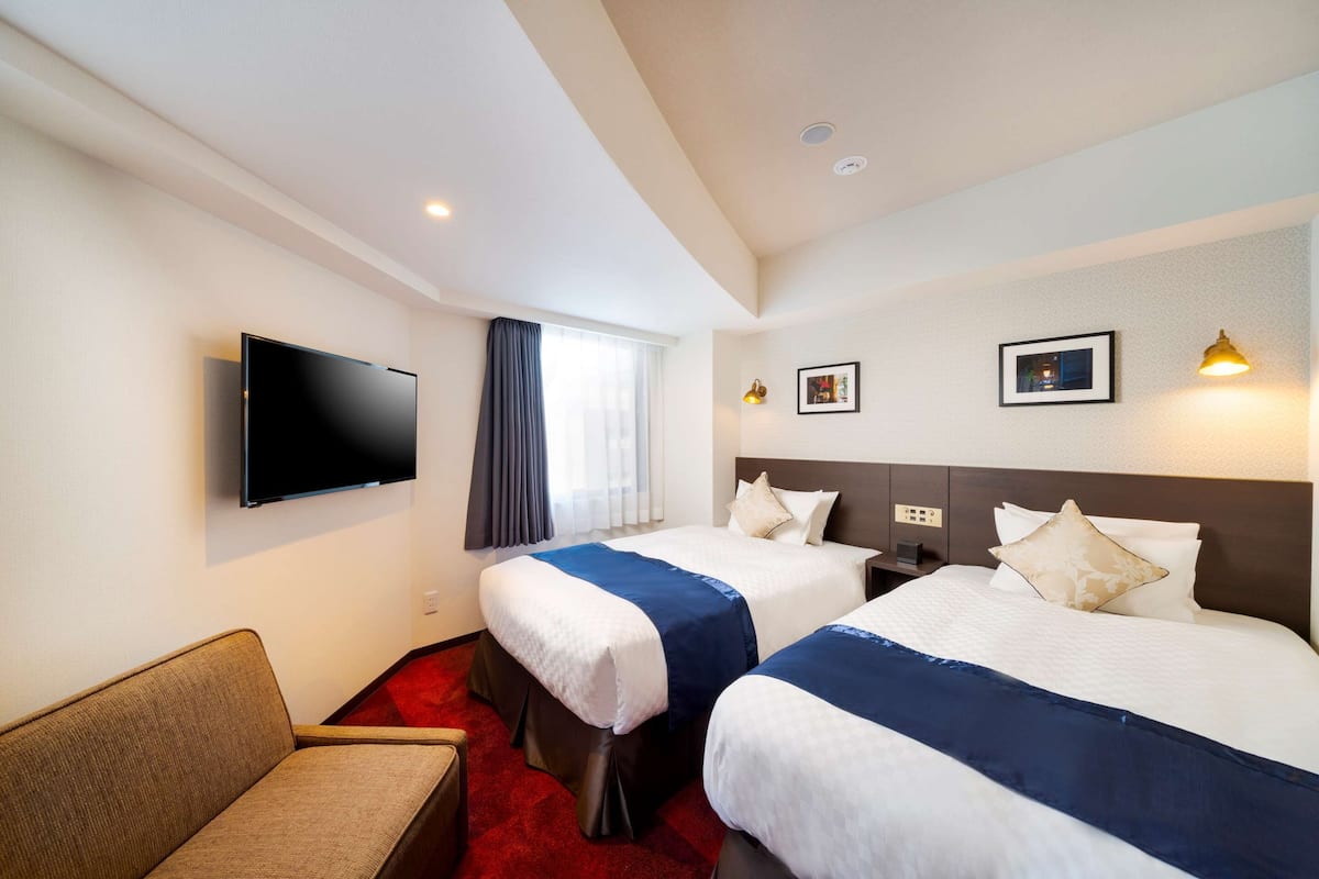 Ein Hotelzimmer mit zwei Betten, einem Flachbildfernseher, einem Sofa und einem fensterbepflanzten Fenster.