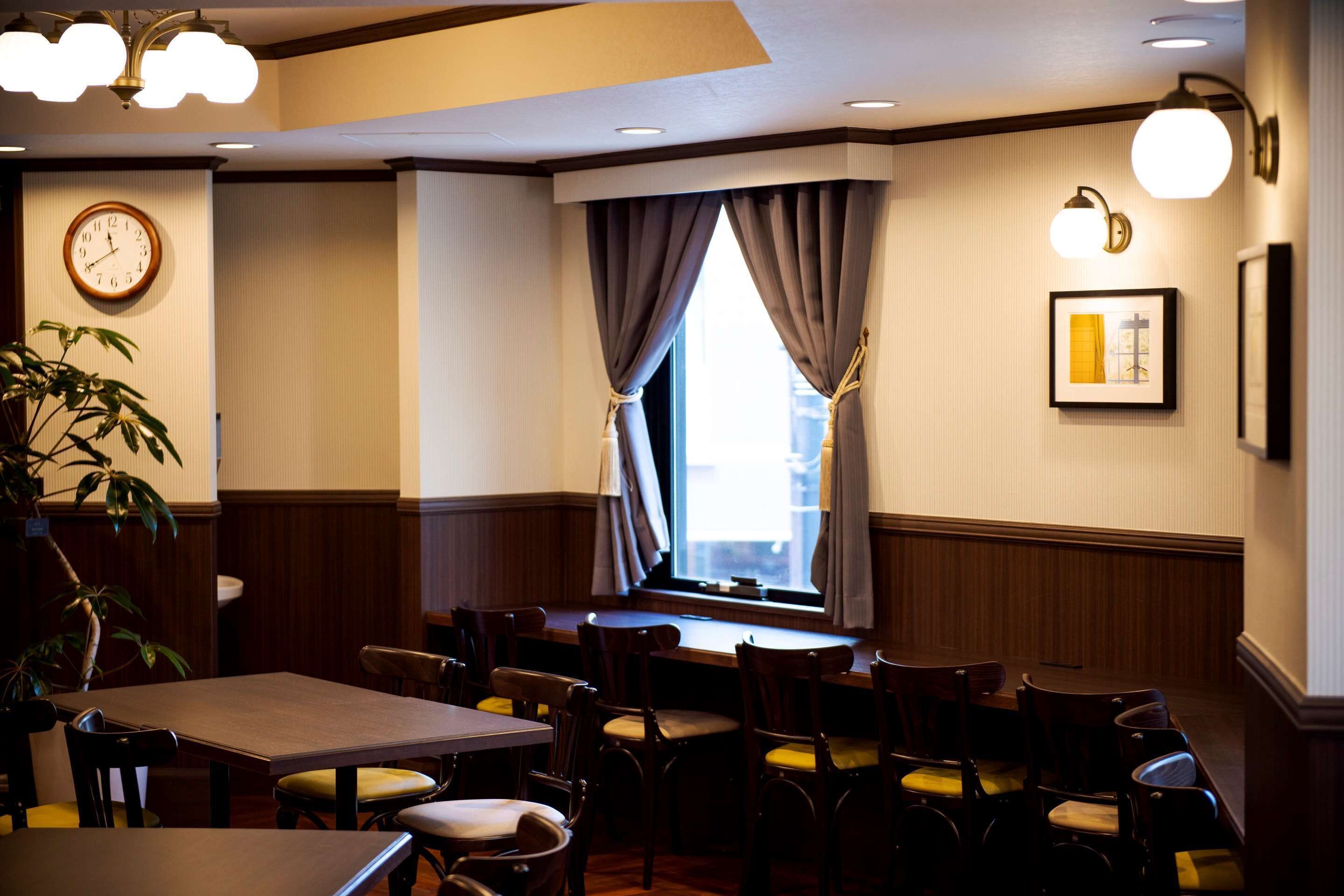 A restaurant with wooden tables and chairs, a clock on the wall, and a window with curtains.