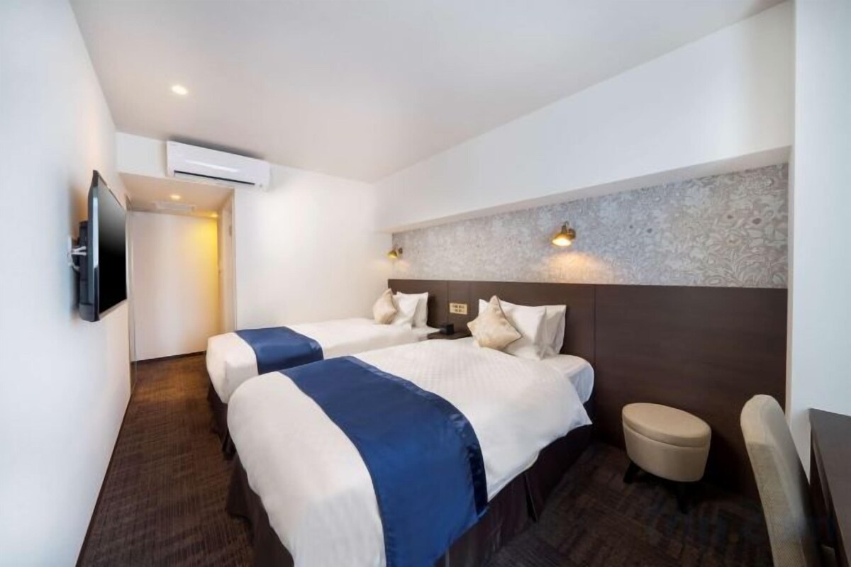 A hotel room with two beds, a television, a small table, and a wall-mounted air conditioning unit.