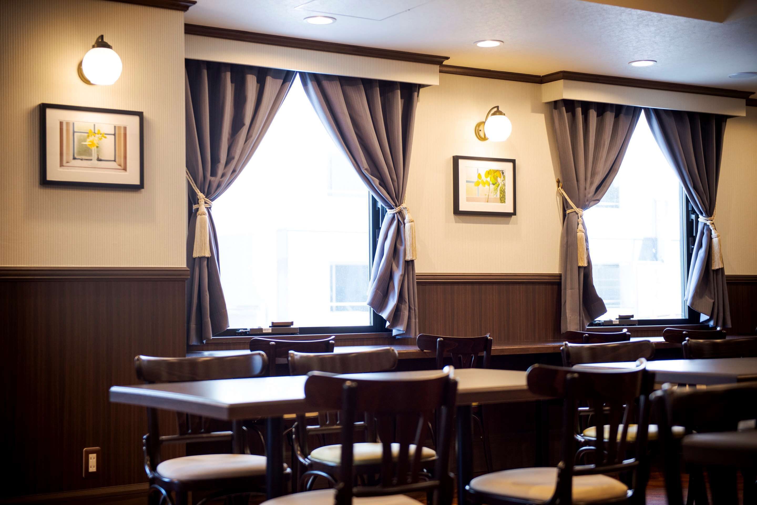 A restaurant with wooden tables and chairs, large windows with curtains, and wall-mounted light fixtures.