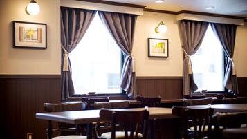 A restaurant with wooden tables and chairs, large windows with curtains, and wall-mounted light fixtures.