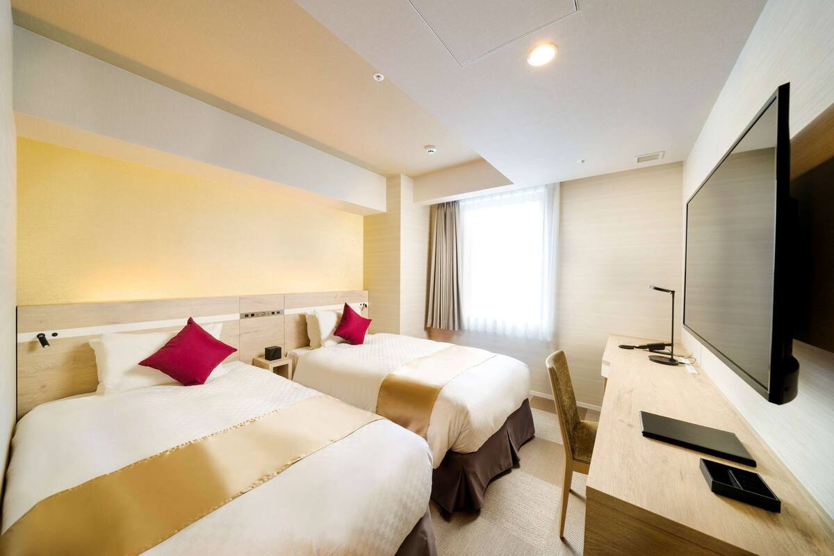 Een hotelkamer met twee bedden, een bureau met een computer, een televisie en een raam met gordijnen.