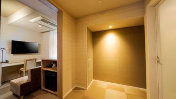 A hotel room with a flat-screen TV, a desk with a chair, a small sofa, a built-in dishwasher, and a closet with a sliding door.