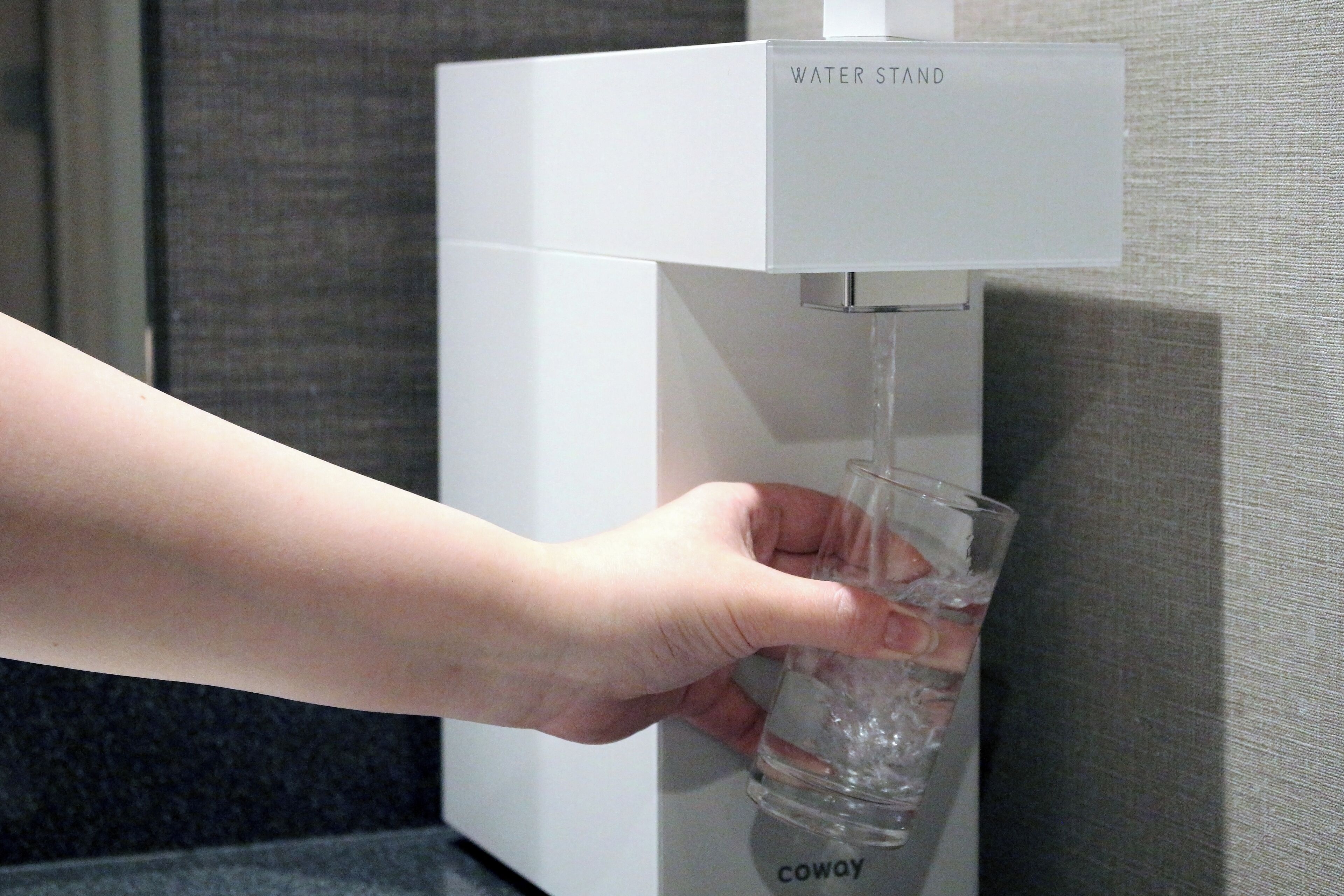 A hand filling a glass from a water dispenser labeled "WATER STAND" with the brand "coway" visible.