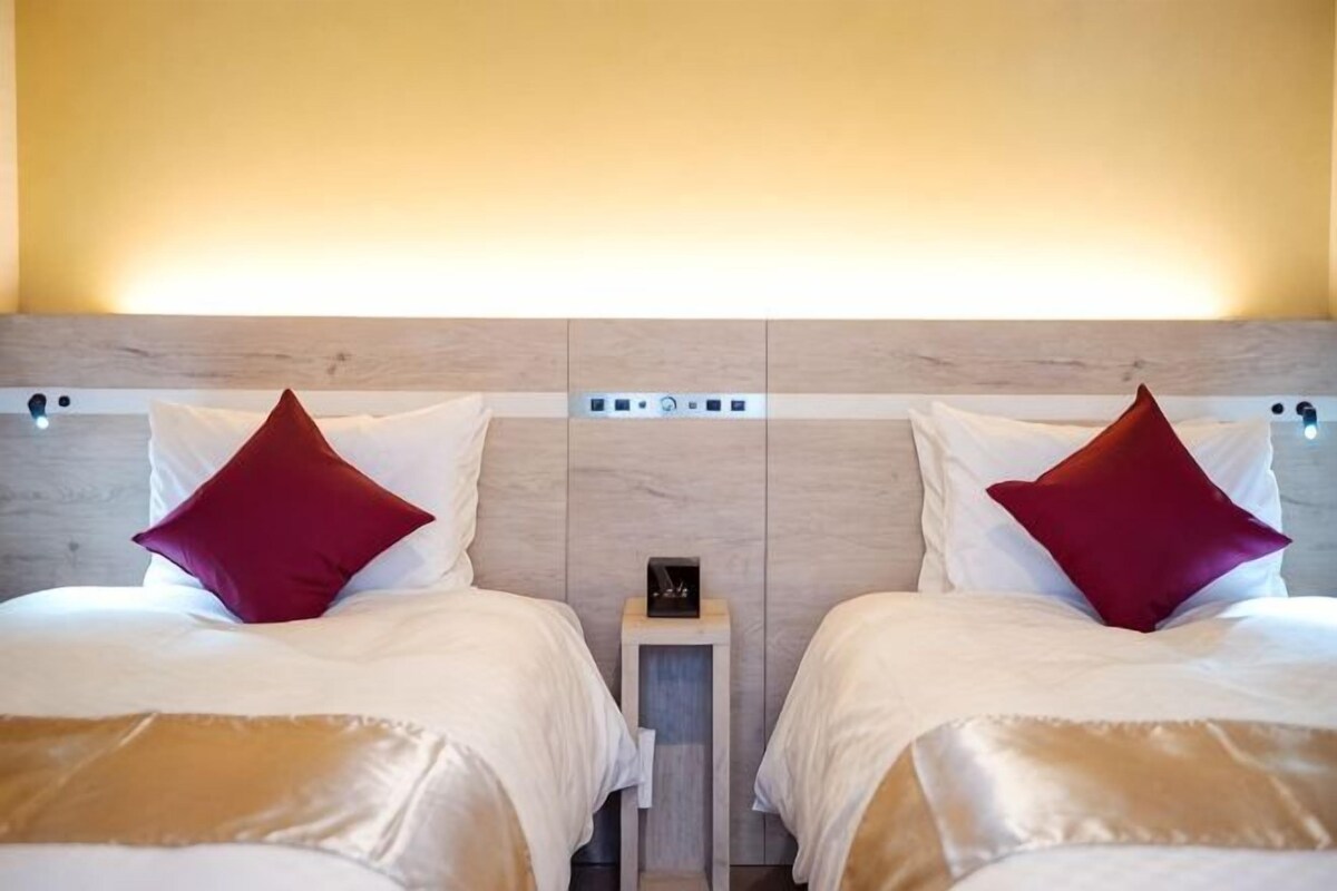 Quarto de hotel com duas camas, cada uma com travesseiro, mesa de cabeceira e luminária na parede.