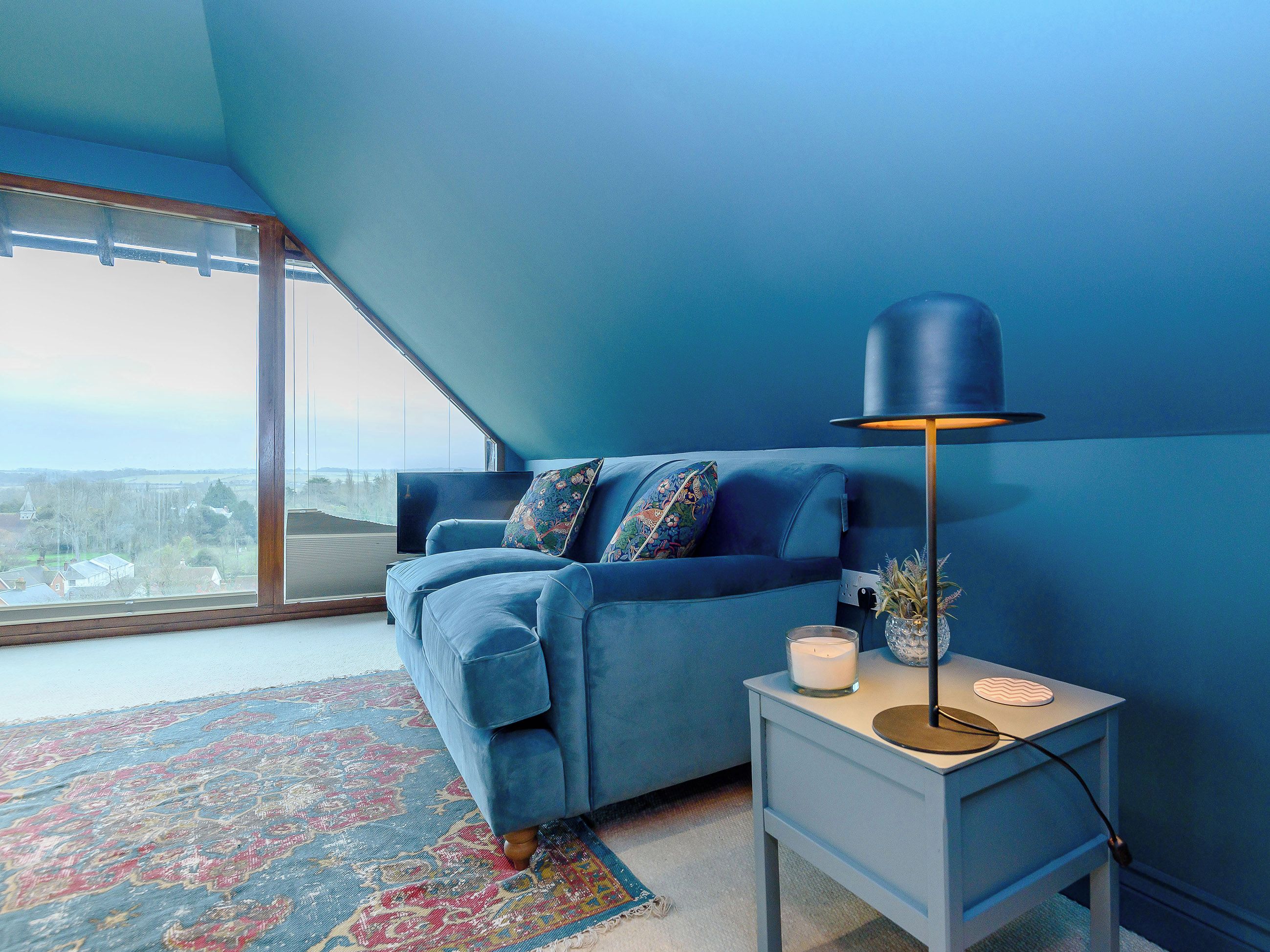 A room with a blue sofa, a small side table with a lamp, and a large window with a view.