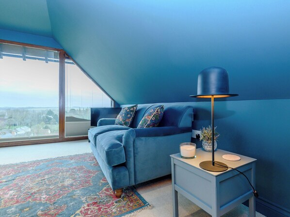 A room with a blue sofa, a small side table with a lamp, and a large window with a view.