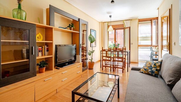 A modern living room with a TV, wooden furniture, a glass coffee table, and a dining area with chairs and a table. - Natural light, quiet, and in a great location (Madrid)