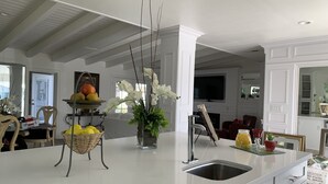 Eine moderne Küche mit einer großen Kochinsel, weißen Schränken und einer integrierten Spüle. Es gibt einen Essbereich mit einem Korbstuhl und einem Obstkorb.