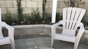 Ein Lagerfeuer mit brennenden Holzscheiten, umgeben von weißen Plastikstühlen, vor einer Steinwand mit Pflanzen.