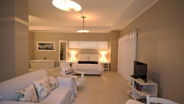 A hotel room with a bed, a sofa, a TV, a bedside table, and a bathroom with a sink and mirror. - Apartment Melkbos Moments-Apartment with a Pool View (Melkbosstrand)