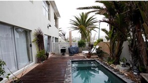 A pool area with a wooden deck, surrounded by potted plants and palm trees. - Apartment Melkbos Moments-Apartment with a Pool View (Melkbosstrand)