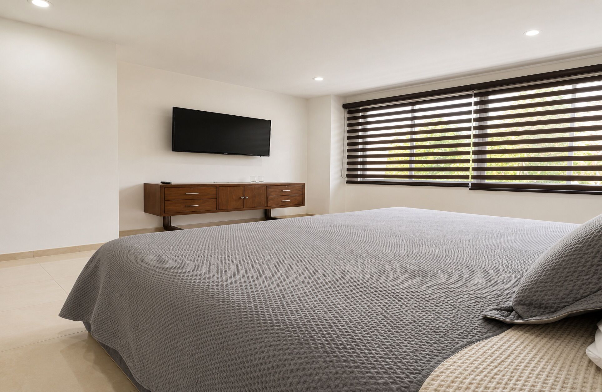 Een slaapkamer met een bed, een aan de muur bevestigde televisie, een raam met zonwering en een houten dressoir.