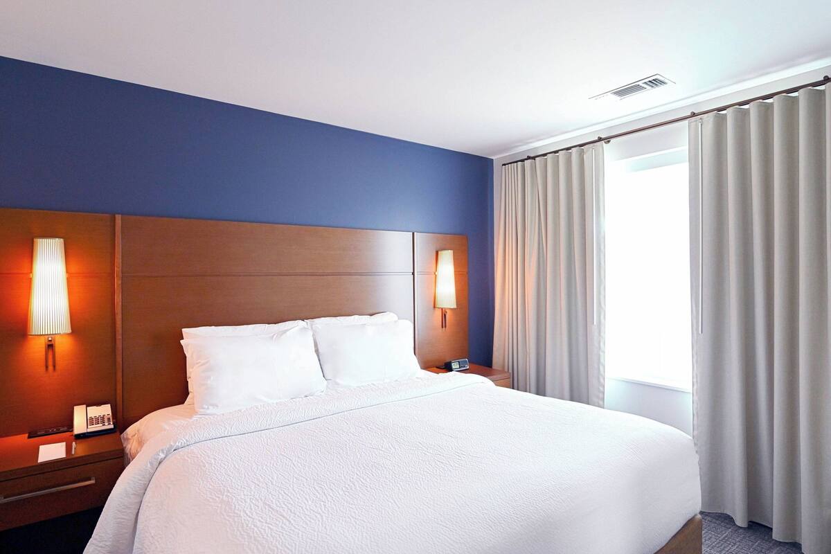 A hotel room with a large bed, two bedside tables, a nightstand, a lamp, and a window with curtains.