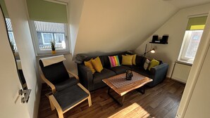 Un salón moderno con sofá, sillón y mesa de centro, con una ventana que ofrece vistas a edificios y un radiador.