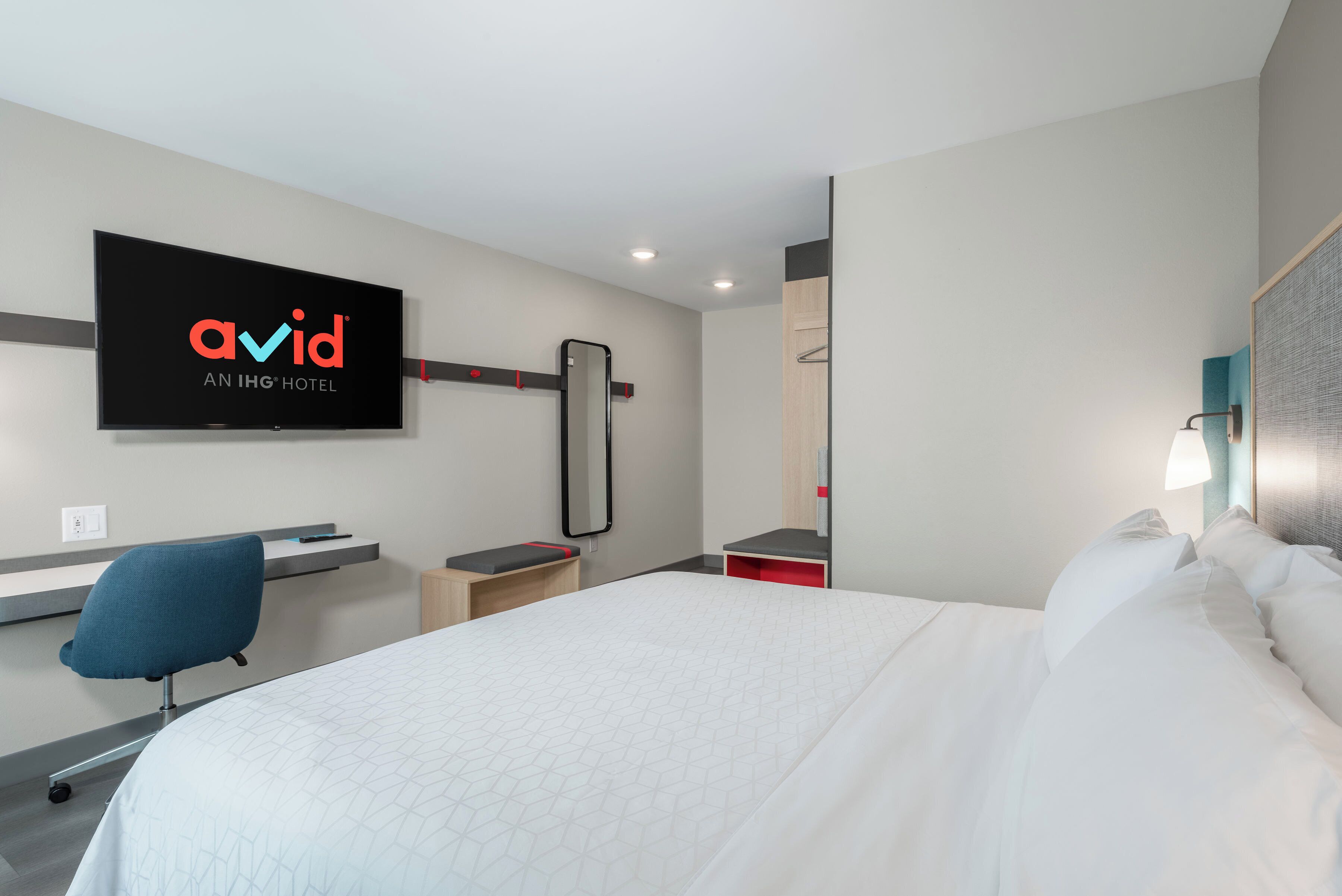 Una habitación de hotel con una cama, un escritorio con silla, un televisor montado en la pared y un espejo.