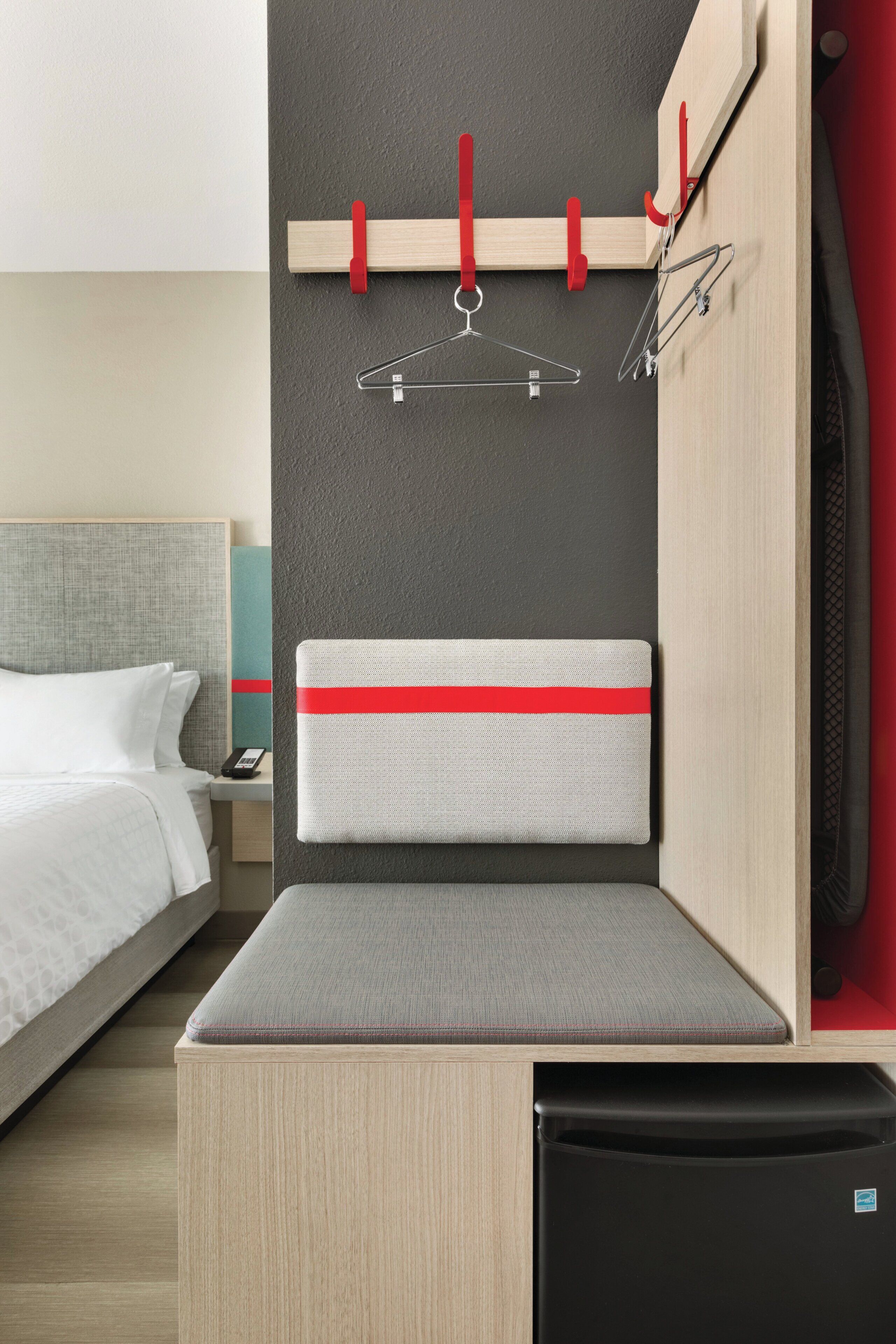 A hotel room with a bed, a bedside table, a nightstand, a red and white striped headboard, a small seating area, and a built-in TV.