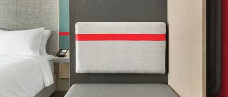 A hotel room with a bed, a bedside table, a nightstand, a red and white striped headboard, a small seating area, and a built-in TV.