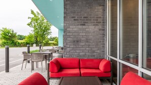 Un canapé rouge et des fauteuils sont placés à l’extérieur d’un bâtiment doté d’un mur en briques et de grandes fenêtres.