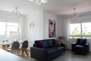 Un salon moderne avec un canapé en cuir noir, une table à manger avec des chaises, un lustre, un ventilateur de plafond et une grande fenêtre donnant sur des immeubles et des arbres.