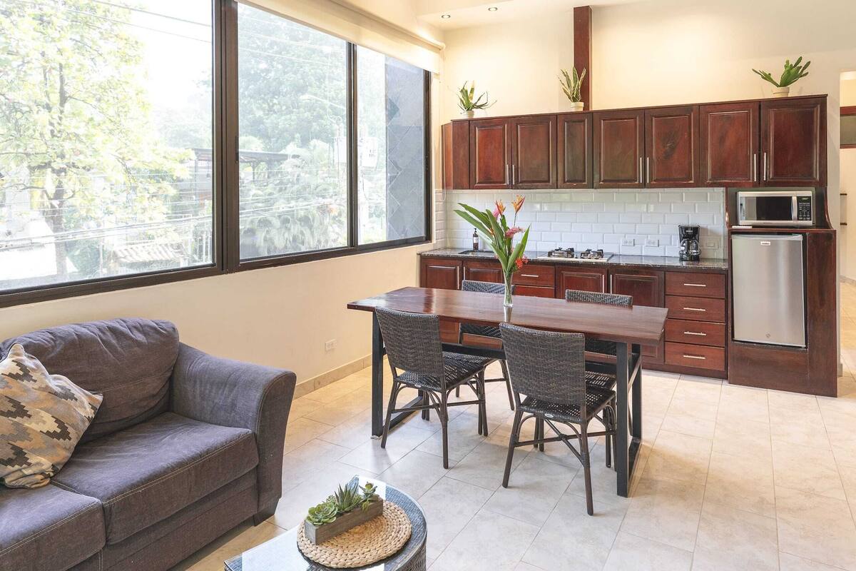A modern kitchen with dark wooden cabinets, a dining table with chairs, a sofa, and a large window offering a view of trees.