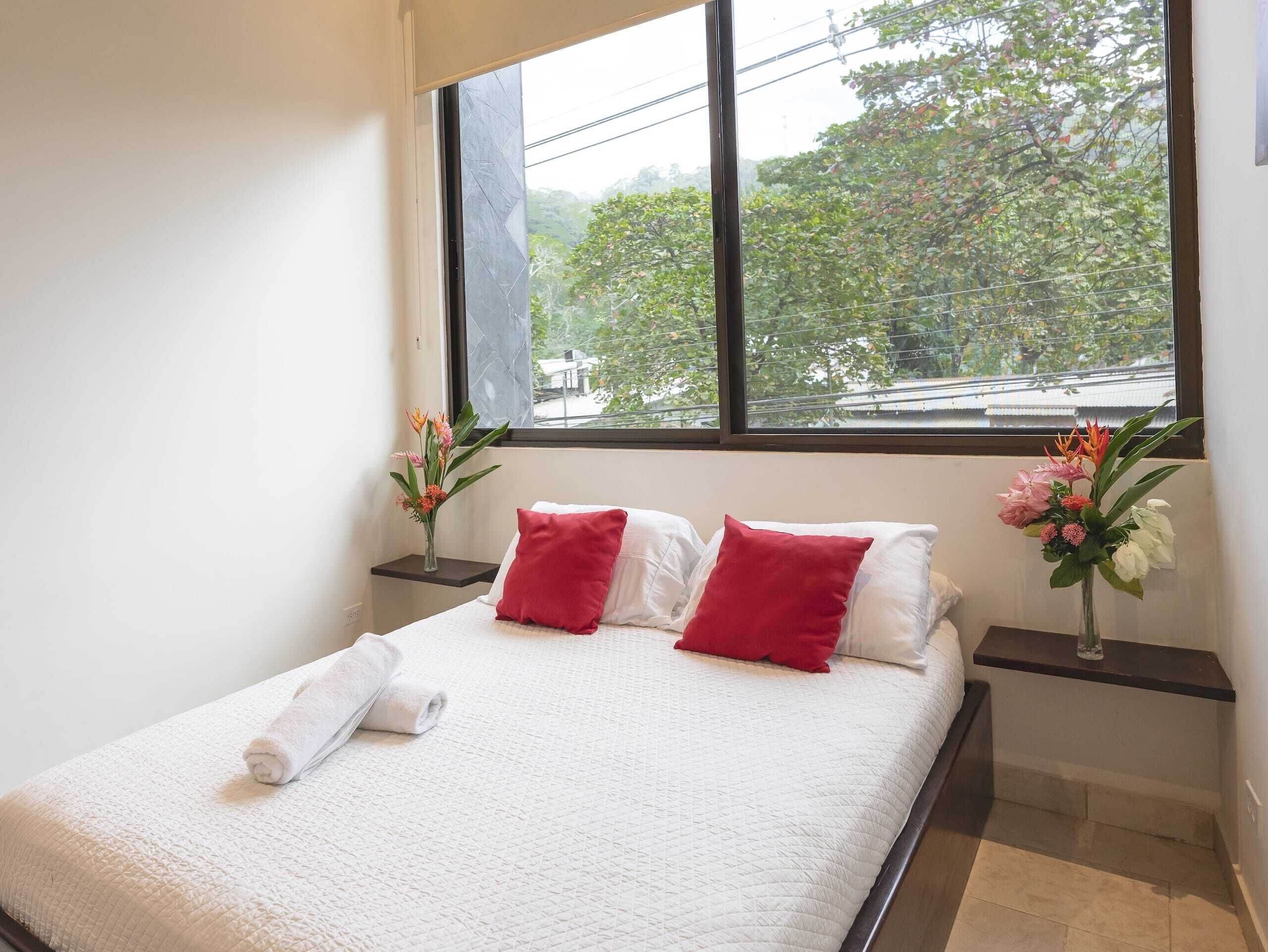 A neatly made bed with white bedding and red pillows, a window with a view of trees, and two bedside tables with flower arrangements.