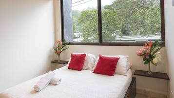 A neatly made bed with white bedding and red pillows, a window with a view of trees, and two bedside tables with flower arrangements.