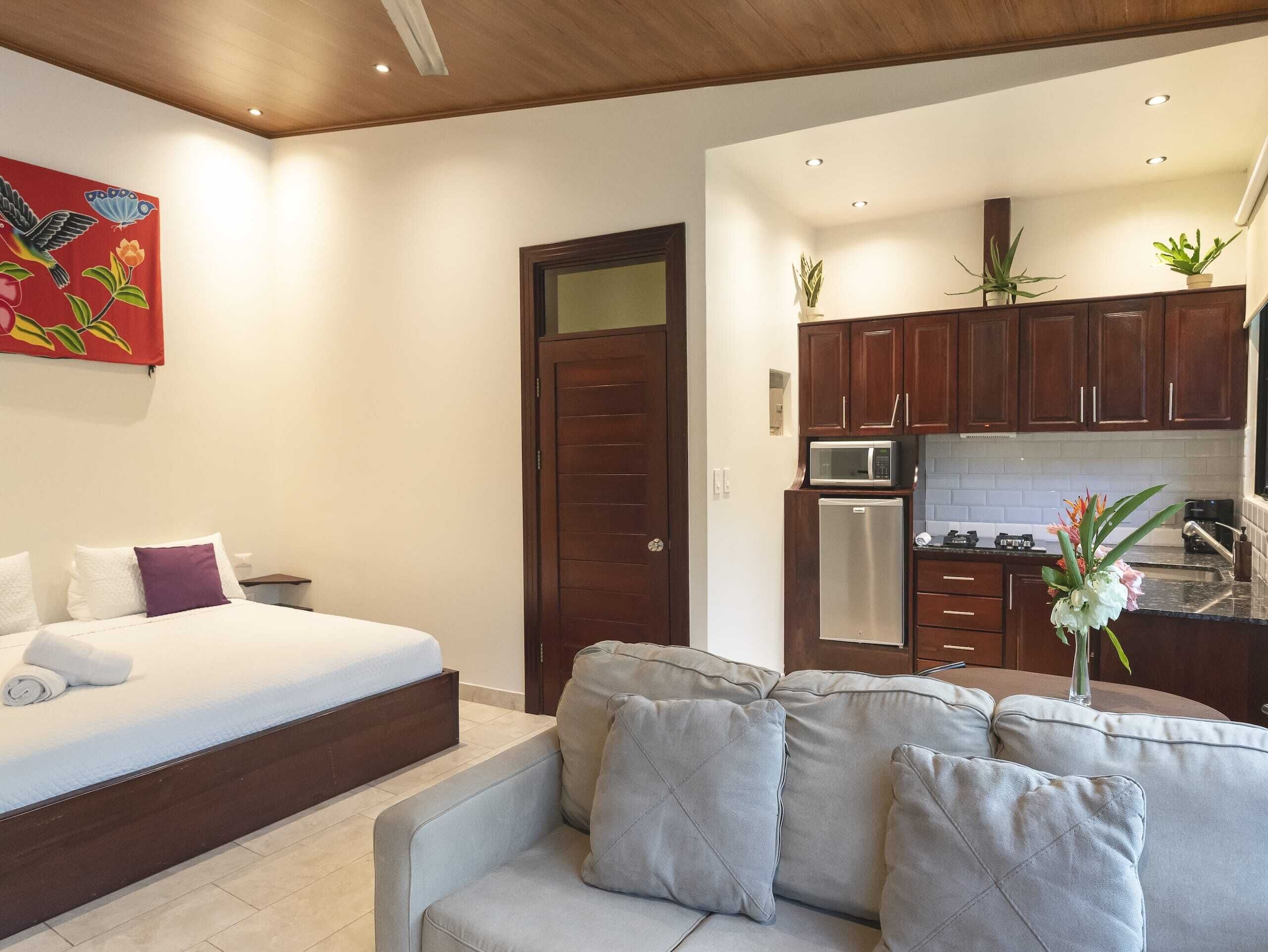 Quarto moderno com cama, sofá cinzento, teto de madeira e cozinha equipada com armários de madeira e micro-ondas.