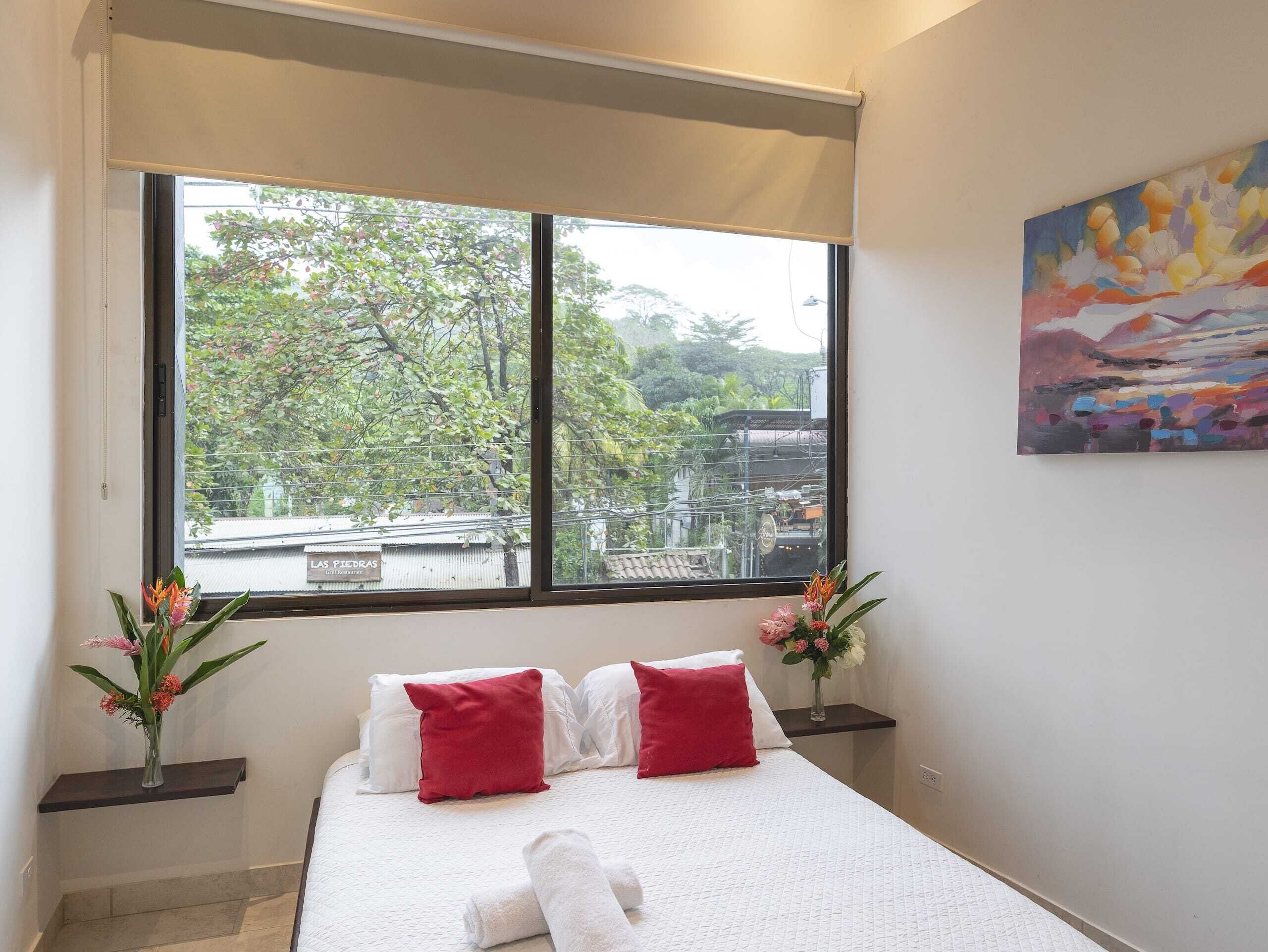 A hotel room with a large window, a bed with white bedding and red pillows, a painting on the wall, and a vase with flowers on a nightstand.