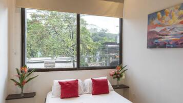 A hotel room with a large window, a bed with white bedding and red pillows, a painting on the wall, and a vase with flowers on a nightstand.